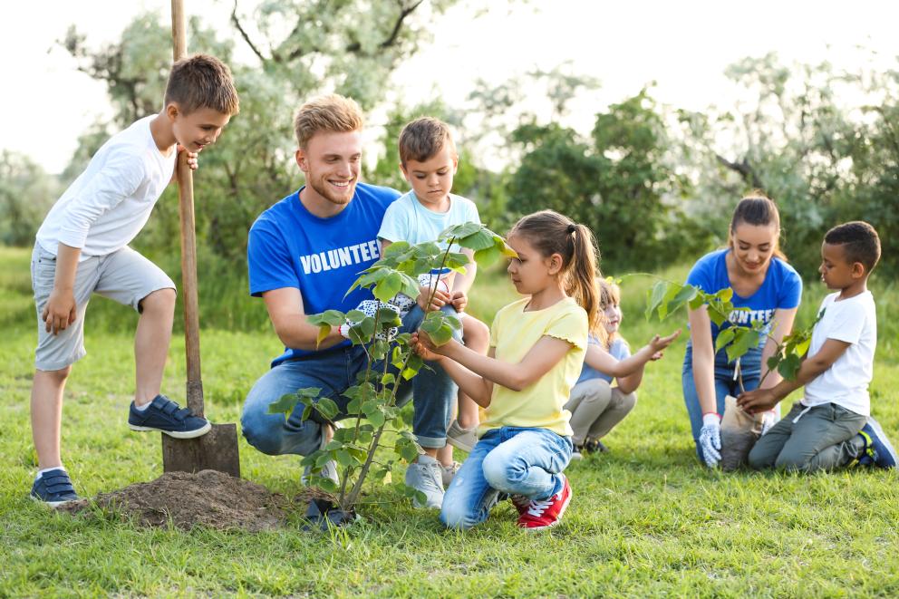 Image shows a male and female volunteer in blue volunteer t-shirts. They are kneeling down to help four young people plant trees. 