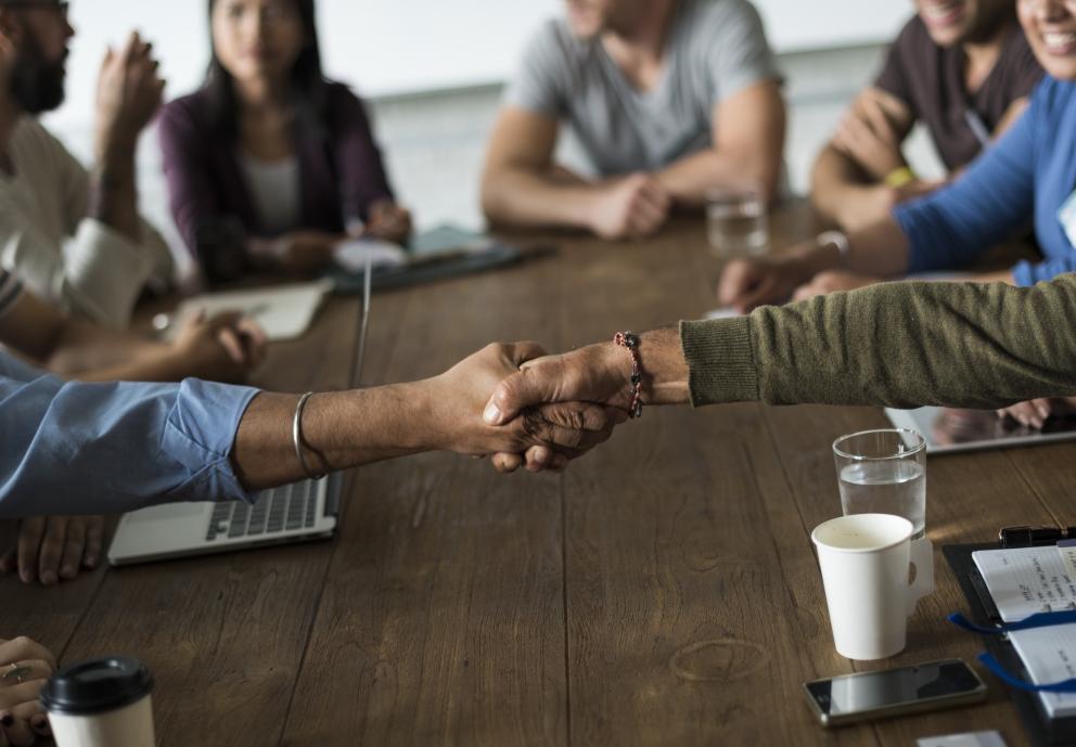 Image shows two people shaking hands in the foreground with people in the background sitting at a desk