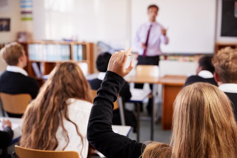 Images shows a class of secondary school pupils at their desks with the teacher at the front of the class. 