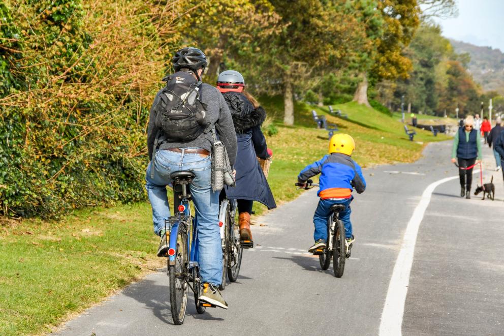 Images shows two adults and a child cycling to the left of the picture with a man walking his dog on the right