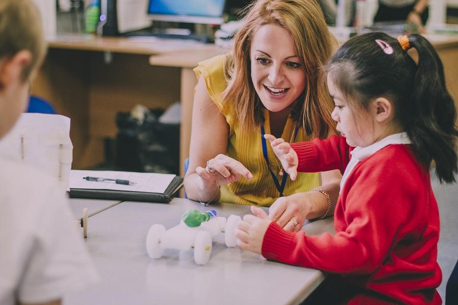 Image shows a young girl sat at a desk with her teacher helping 