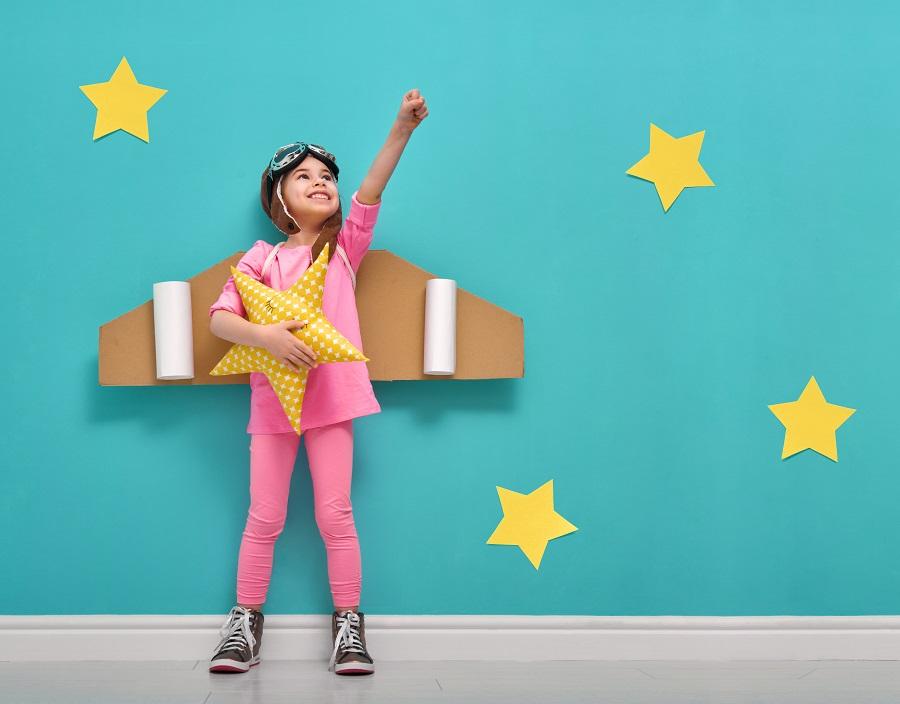 Images shows a girl dressed up in pink with cardboard airplane wings against a blue background with yellow stars