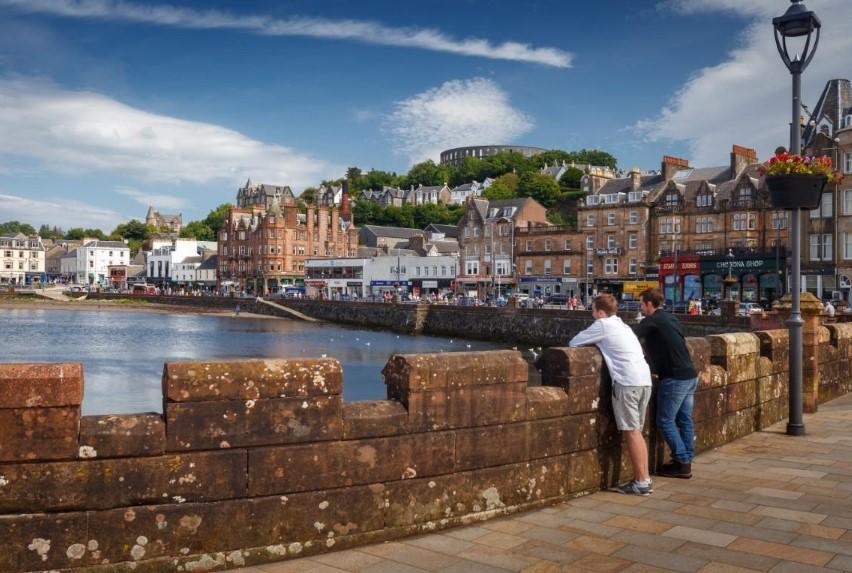  Image shows two visitors leaning on the wall at Oban harbour looking out to waterout to