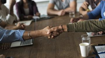 Image shows two people shaking hands in the foreground with people in the background sitting at a desk