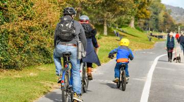 Images shows two adults and a child cycling to the left of the picture with a man walking his dog on the right