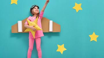 Images shows a girl dressed up in pink with cardboard airplane wings against a blue background with yellow stars
