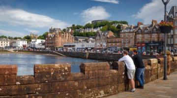  Image shows two visitors leaning on the wall at Oban harbour looking out to waterfront.