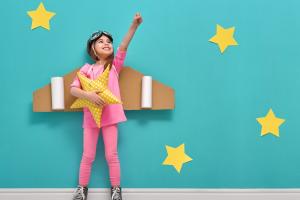 Images shows a girl dressed up in pink with cardboard airplane wings against a blue background with yellow stars